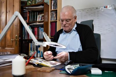 Billede af en ældre mand som sidder i stolen og kigger i gennem en stor lup med en bog på bordet