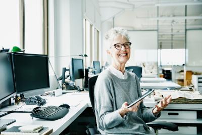 Ældre dame med skærmbriller på, som sidder på kontoret og smiler med en IPad i hånden