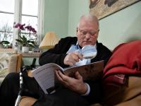 Billede af en ældre mand som sidder i en sofa med en lup i hånden og læser et blad