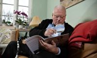 Billede af en ældre mand som sidder i en sofa med en lup i hånden og læser et blad